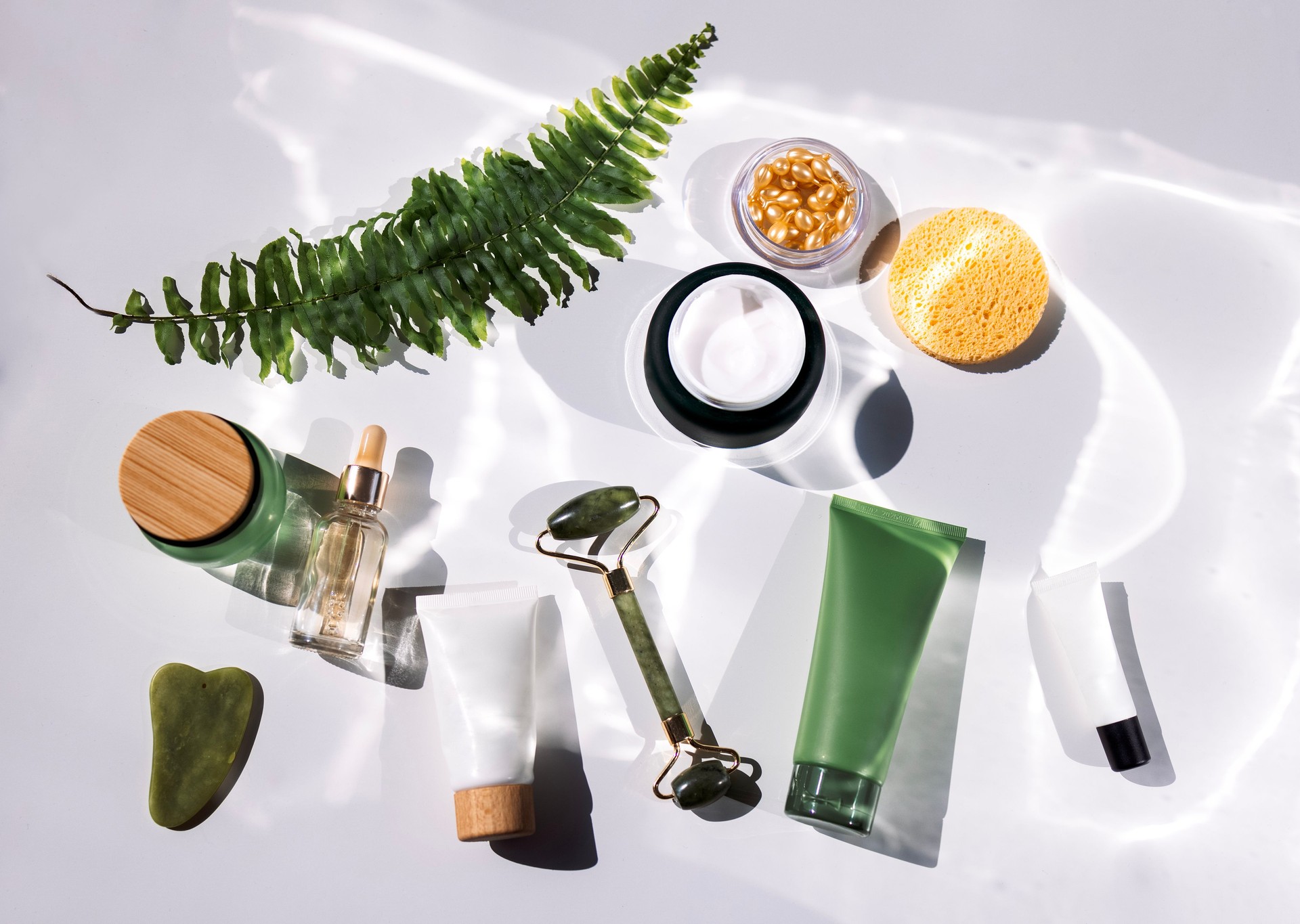 A set of mockups of ecological cosmetics with a fern leaf. Various cream tubes, a bottle of serum, capsules, a sponge, a jade facial massager and gouache scraper.